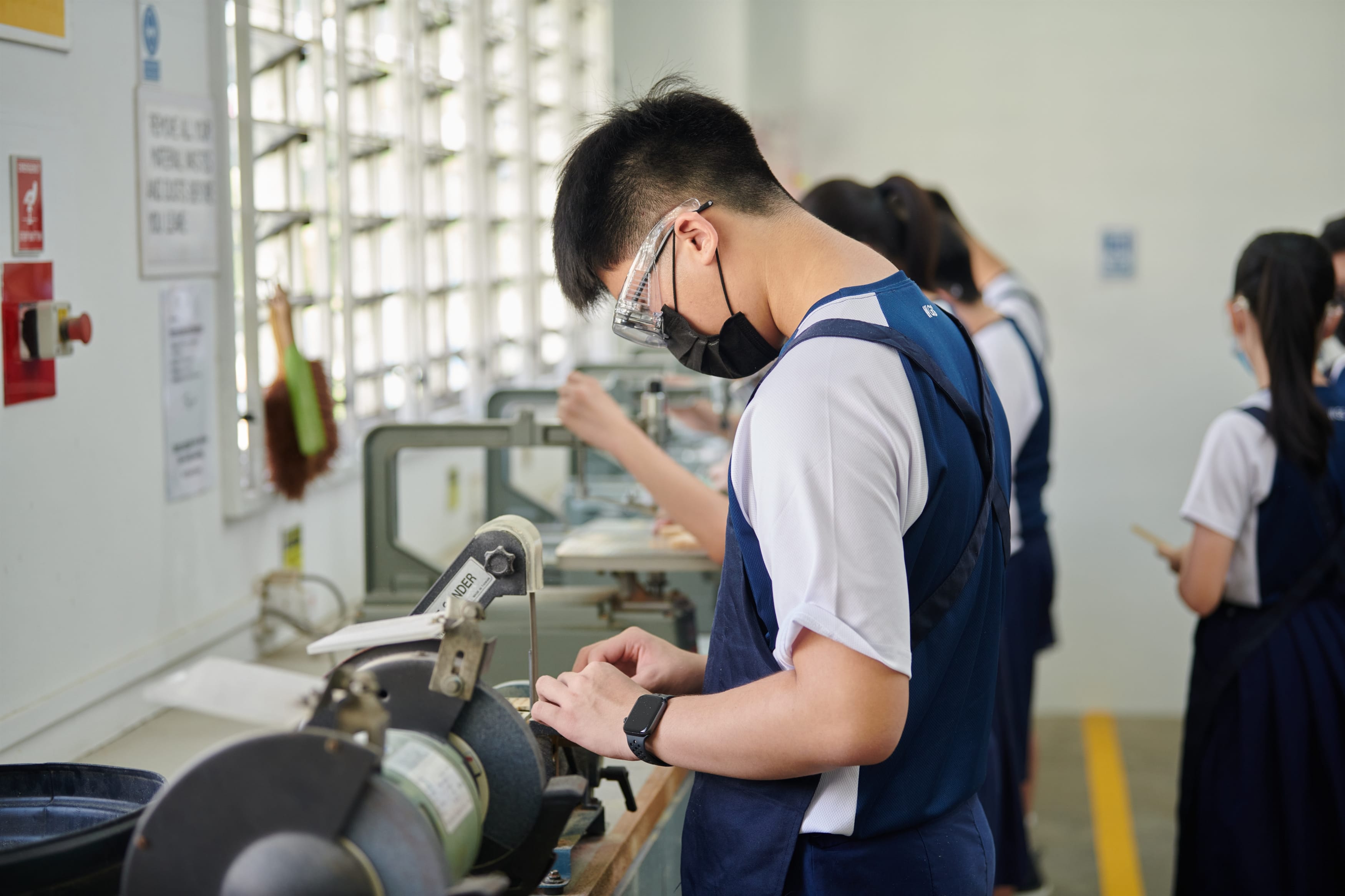 A student wearing safety goggles and a face mask operates a bench grinder in a workshop class.